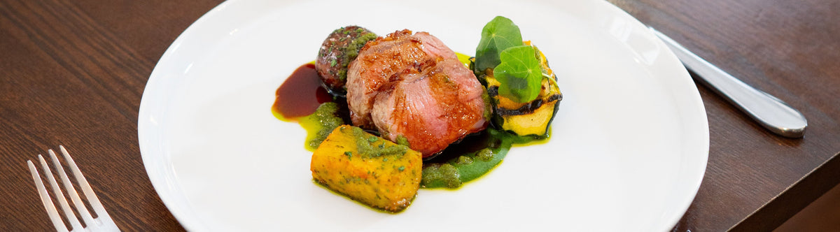A plated gourmet dish of lamb with green beans and roasted vegetables on a white plate, set on a dark wooden dining table next to a glass of red wine, a small vase of flowers, and silverware.