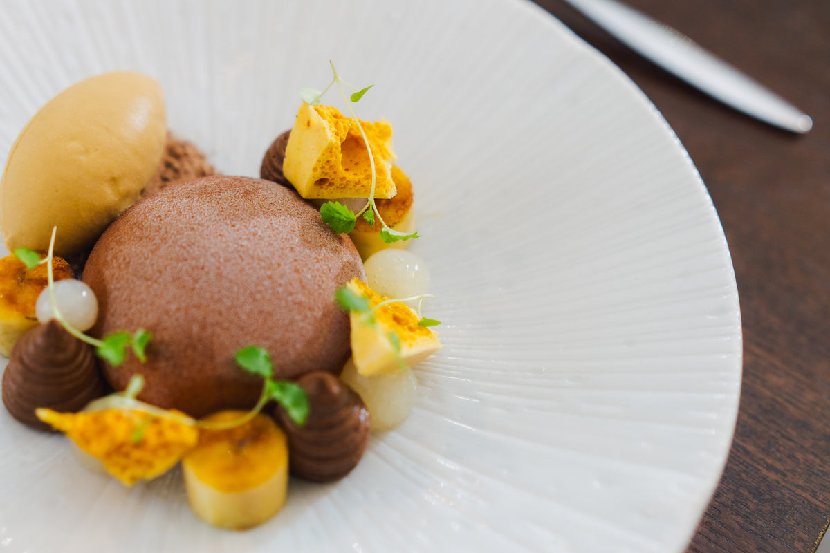 A close-up of a gourmet dessert plated on a white ribbed ceramic dish, featuring a scoop of chocolate parfait, a ice cream, orange-yellow fruit pieces, and microgreens on a dark wooden table.